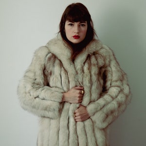 Op de afbeelding: Een vrouw die een witte bontjas draagt met lang, stijl haar. Ze kijkt recht in de camera met een serieuze uitdrukking.