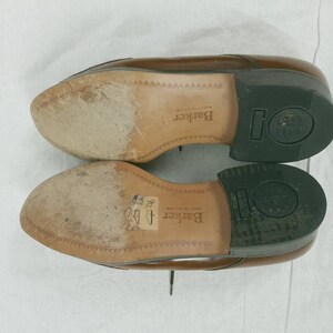 May include: Close-up of the soles of two brown leather shoes. The shoes are made by Barker and have a black rubber sole. The soles are worn and have a textured pattern.