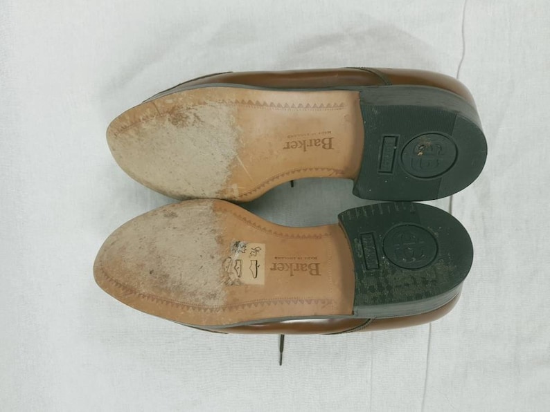May include: Close-up of the soles of two brown leather shoes. The shoes are made by Barker and have a black rubber sole. The soles are worn and have a textured pattern.