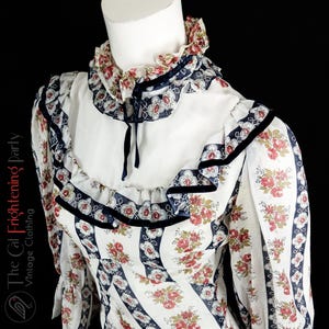 May include: A white vintage dress with a floral print and a ruffled collar. The dress has a navy blue trim and a black velvet ribbon.