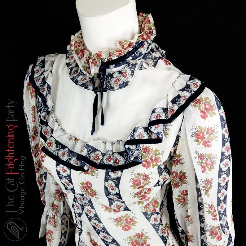 May include: A white vintage dress with a floral print and a ruffled collar. The dress has a navy blue trim and a black velvet ribbon.