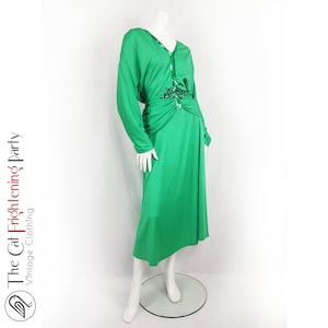 Puede incluir: Un vestido largo de manga larga, de color verde esmeralda, con cintura fruncida y un adorno de lentejuelas. El vestido tiene un escote en V y una falda fluida.