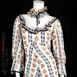May include: A white vintage dress with a floral print and blue stripes. The dress has a high neck with a ruffle collar and long sleeves with ruffles at the cuffs.