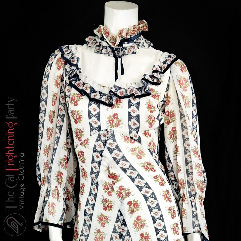 May include: A white vintage dress with a floral print and blue stripes. The dress has a high neck with a ruffle collar and long sleeves with ruffles at the cuffs.