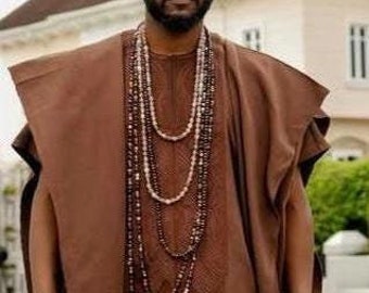 May include: A man wearing a brown long-sleeved shirt with a brown patterned design, brown pants, and black shoes. He is wearing a long brown necklace with multiple strands of beads. He is holding a black walking stick.