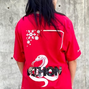 Puede incluir: Camiseta roja con un diseño de serpiente blanca y la palabra "snow" en una fuente estilizada. La camiseta también presenta gráficos de copos de nieve blancos. El modelo lleva vaqueros azules.