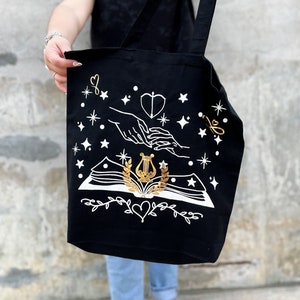 May include: Black tote bag with a white design of two hands holding a book with a lyre and laurel wreath. The design is surrounded by stars and hearts.