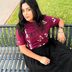 Peut inclure: Une femme portant un t-shirt bordeaux avec des papillons dor&eacute;s et le texte "Vampire pr&eacute;f&eacute;r&eacute;" imprim&eacute; dessus. Elle porte &eacute;galement une jupe en tulle noire et est assise sur un banc en m&eacute;tal noir.