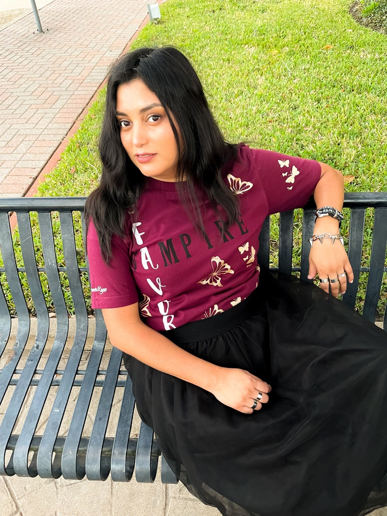 Peut inclure: Une femme portant un t-shirt bordeaux avec des papillons dor&eacute;s et le texte "Vampire pr&eacute;f&eacute;r&eacute;" imprim&eacute; dessus. Elle porte &eacute;galement une jupe en tulle noire et est assise sur un banc en m&eacute;tal noir.