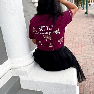 Peut inclure: Un t-shirt bordeaux avec des motifs de papillons dor&eacute;s et le texte "NCT 127 You'll always be my favorite".