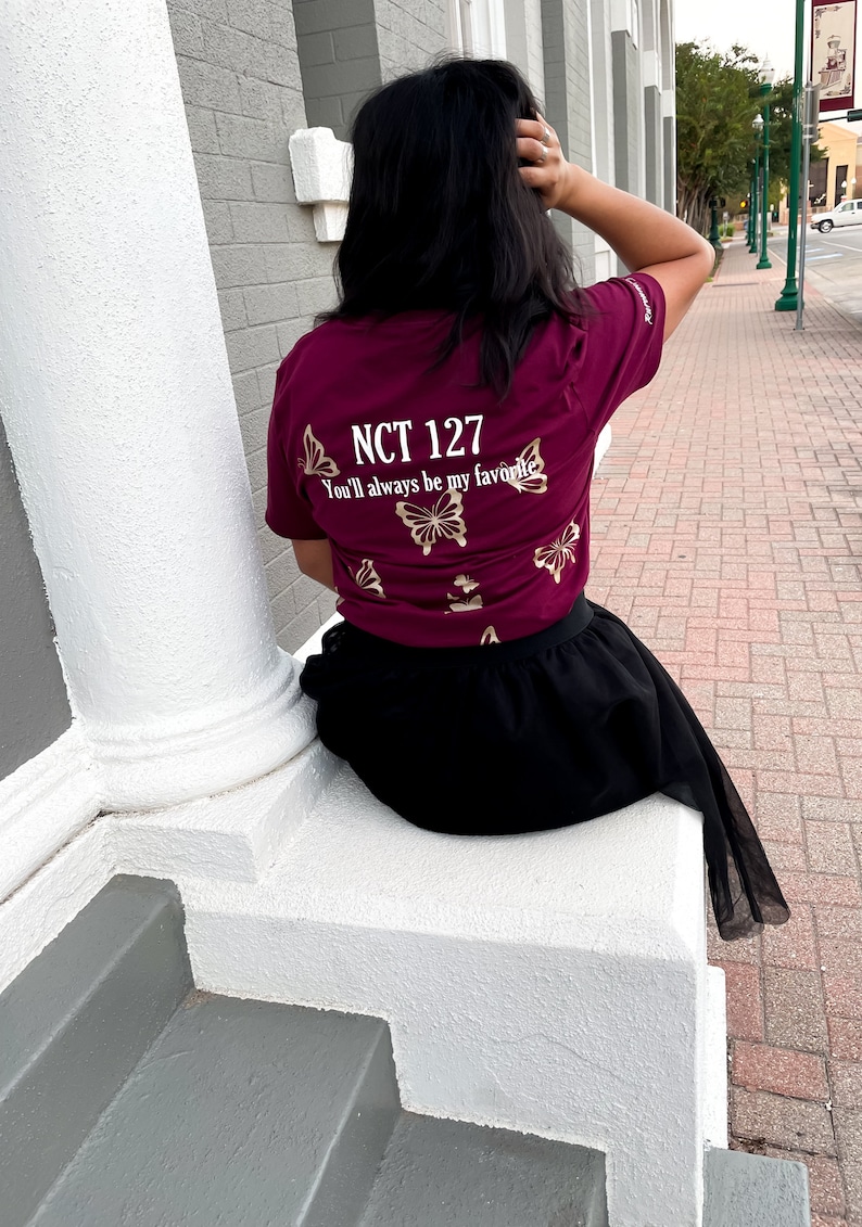 Peut inclure: Un t-shirt bordeaux avec des motifs de papillons dor&eacute;s et le texte "NCT 127 You'll always be my favorite".