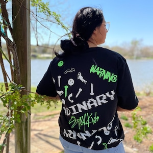 Puede incluir: Una camiseta negra con un estampado gráfico verde y blanco. El gráfico incluye las palabras "WARNING" y "BINARY" y una variedad de imágenes pequeñas, incluyendo tornillos, pájaros y flechas. El texto "Stay4Kids" también está incluido en el gráfico.