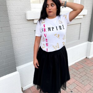 Peut inclure: T-shirt blanc avec les mots "FAVORITE VAMPIRE" imprim&eacute;s verticalement en noir et rose. Le t-shirt est d&eacute;cor&eacute; de motifs de papillons dor&eacute;s.