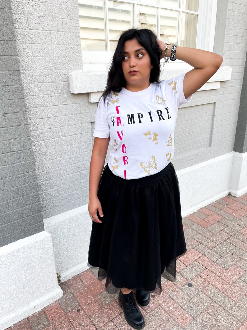 Peut inclure: T-shirt blanc avec les mots "FAVORITE VAMPIRE" imprim&eacute;s verticalement en noir et rose. Le t-shirt est d&eacute;cor&eacute; de motifs de papillons dor&eacute;s.