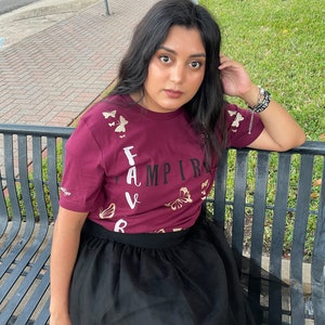 Peut inclure: Une personne portant un t-shirt bordeaux avec des papillons dor&eacute;s et le mot "FAVAMPIRE" imprim&eacute; dessus. Elle porte une jupe en tulle noire et est assise sur un banc en m&eacute;tal noir.