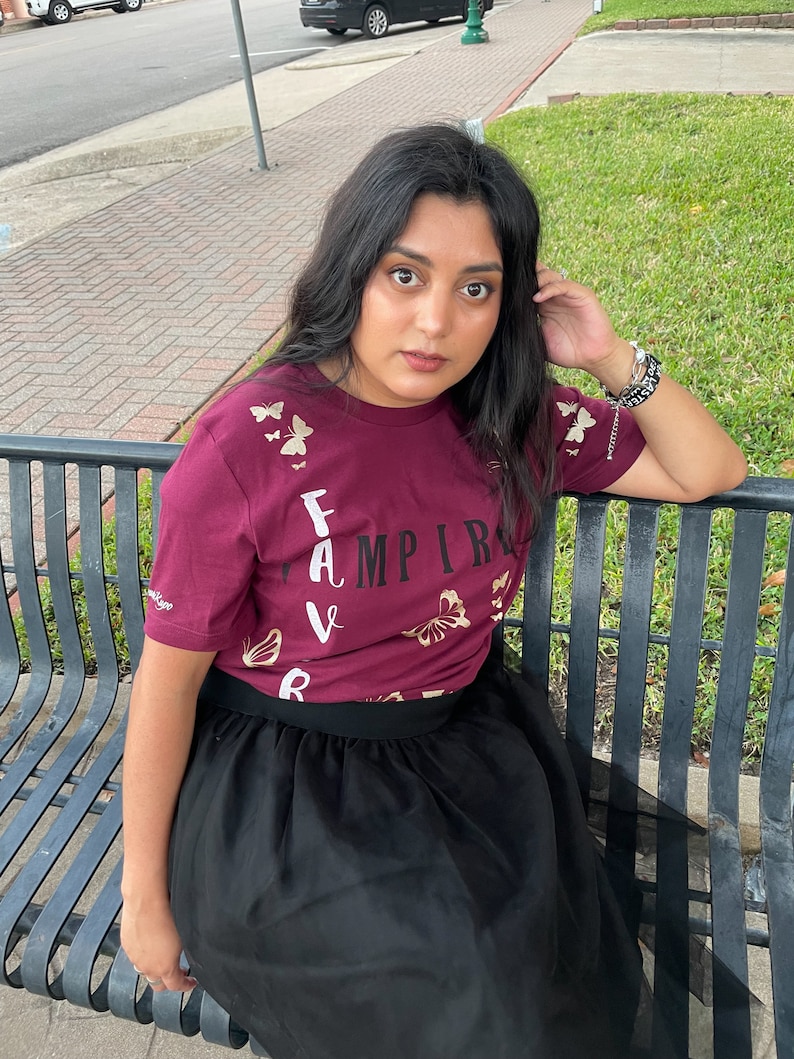 Peut inclure: Une personne portant un t-shirt bordeaux avec des papillons dor&eacute;s et le mot "FAVAMPIRE" imprim&eacute; dessus. Elle porte une jupe en tulle noire et est assise sur un banc en m&eacute;tal noir.