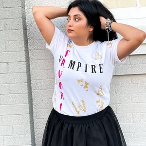 Peut inclure: T-shirt blanc avec le texte "Vampire pr&eacute;f&eacute;r&eacute;" imprim&eacute; verticalement en noir et rose. Le t-shirt a un motif de papillon dor&eacute;.