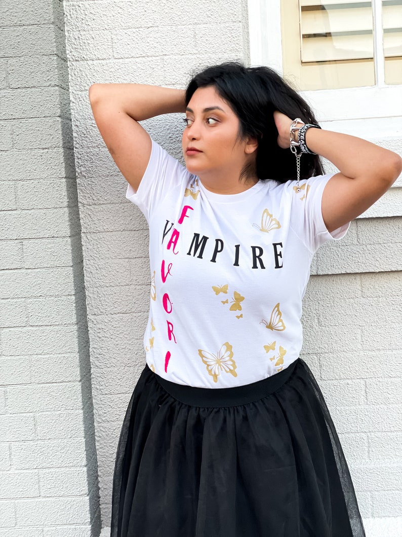 Peut inclure: T-shirt blanc avec le texte "Vampire pr&eacute;f&eacute;r&eacute;" imprim&eacute; verticalement en noir et rose. Le t-shirt a un motif de papillon dor&eacute;.