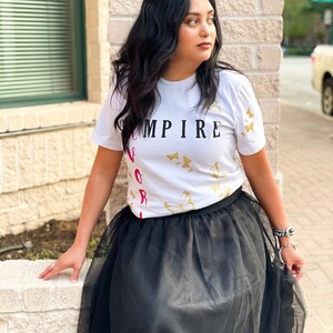Peut inclure: Une femme portant un t-shirt blanc avec le texte "EMPIRE" et des motifs de papillons dor&eacute;s. Elle porte &eacute;galement une jupe en tulle noire.