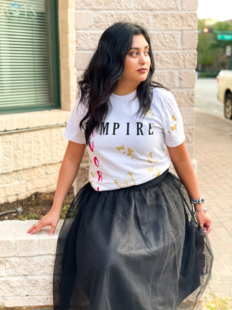Peut inclure: Une femme portant un t-shirt blanc avec le texte "EMPIRE" et des motifs de papillons dor&eacute;s. Elle porte &eacute;galement une jupe en tulle noire.