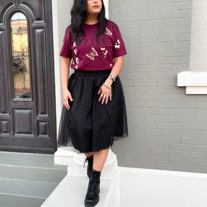 Peut inclure: Une femme portant un t-shirt bordeaux avec des papillons dor&eacute;s et le mot "EMPIRE" imprim&eacute; dessus. Elle porte &eacute;galement une jupe en tulle noire et des bottes de combat noires.