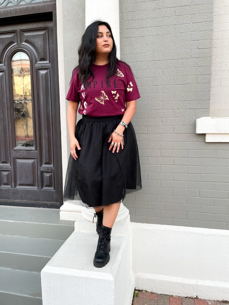 Peut inclure: Une femme portant un t-shirt bordeaux avec des papillons dor&eacute;s et le mot "EMPIRE" imprim&eacute; dessus. Elle porte &eacute;galement une jupe en tulle noire et des bottes de combat noires.