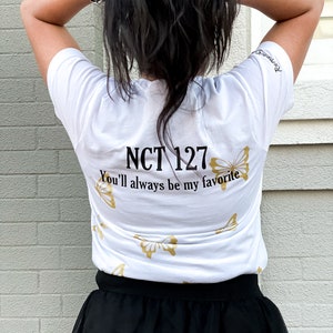Peut inclure: T-shirt blanc avec du texte dor&eacute; qui dit "NCT 127 You'll always be my favorite" et des motifs de papillons dor&eacute;s. Le t-shirt est port&eacute; par une personne aux cheveux longs et noirs.