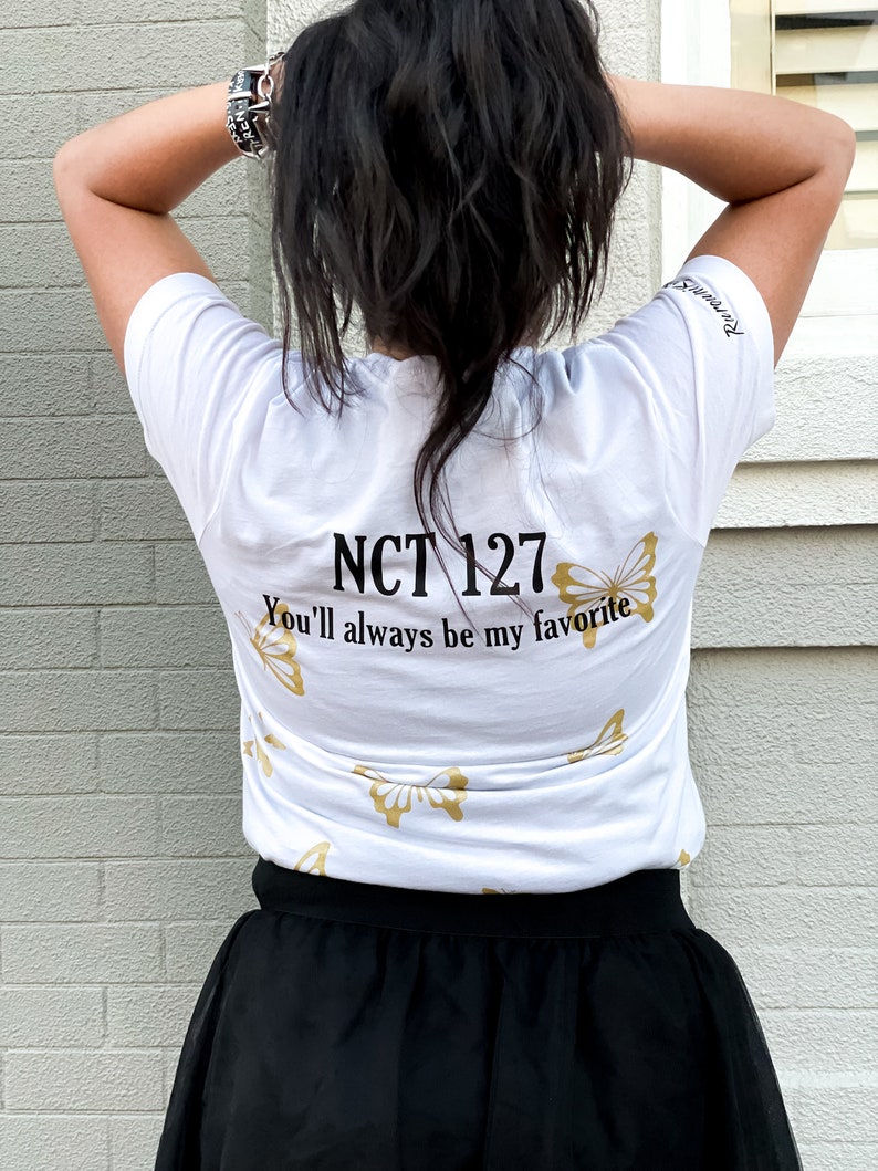 Peut inclure: T-shirt blanc avec du texte dor&eacute; qui dit "NCT 127 You'll always be my favorite" et des motifs de papillons dor&eacute;s. Le t-shirt est port&eacute; par une personne aux cheveux longs et noirs.