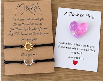 May include: Two black cord bracelets on a tan card featuring a gold sun charm and a silver moon charm with text about friendship. Beside it, a white card with a pink and purple glitter heart charm and the text A Pocket Hug.