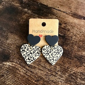 May include: A pair of black and white heart-shaped earrings with a leopard print design. The earrings are handmade and have a black heart on top of a white heart with a leopard print.
