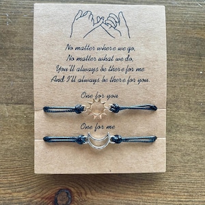 May include: Two matching friendship bracelets with black cords and metal charms, one a sun and the other a crescent moon, presented on a brown card. The card features the text "No matter where we go..." and "One for you, One for me."