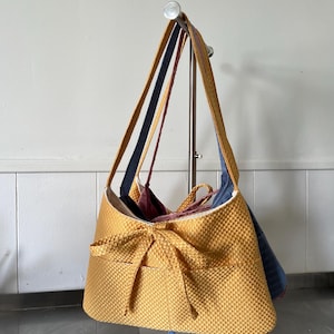 May include: A mustard yellow shoulder bag with a textured waffle-weave fabric and a decorative bow detail on the front. The bag features long shoulder straps and is displayed alongside other bags in shades of navy blue and deep red.