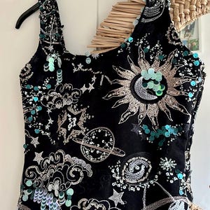 Könnte beinhalten: Schwarzes Tanktop mit einem Himmelsdesign mit Sonne, Mond, Sternen und Planeten. Das Oberteil ist mit silberner Stickerei und türkisfarbenen Pailletten verziert. Das Tanktop hängt an einem schwarzen Kleiderbügel.