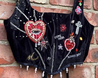 May include: Black velvet corset top with elaborate embellishments. Features beaded heart and eye designs, crosses, stars, and the phrases "COME HERE" and "RIGHT NOW". Includes pearl and crystal accents, and black straps.