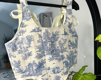 May include: A white linen corset top with a blue toile pattern featuring a pastoral scene. The corset has a sweetheart neckline and adjustable straps with satin ribbon ties.
