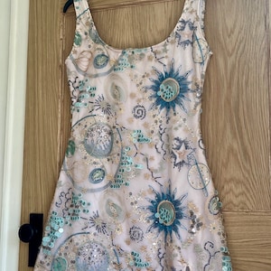 以下が含まれることがあります： ピンクのベースに、青と緑のビーズと刺繍で模様が施された、袖なしのミニドレス。模様には、円形、星形、その他の抽象的な形が含まれています。