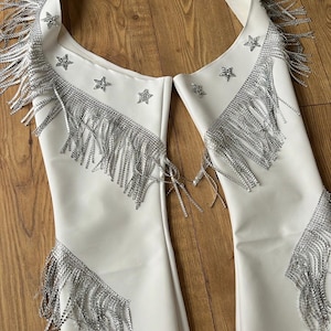 Puede incluir: Chaleco blanco con flecos plateados y detalles de estrellas. El chaleco presenta un diseño de cuello halter con paneles largos. Los flecos plateados están adheridos al escote, los hombros y los paneles inferiores. El chaleco es de material liso y opaco.
