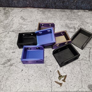 May include: Six small, 3D printed boxes in various colors, including black, purple, and gold. The boxes are stacked on top of each other. There are four small screws on the surface.