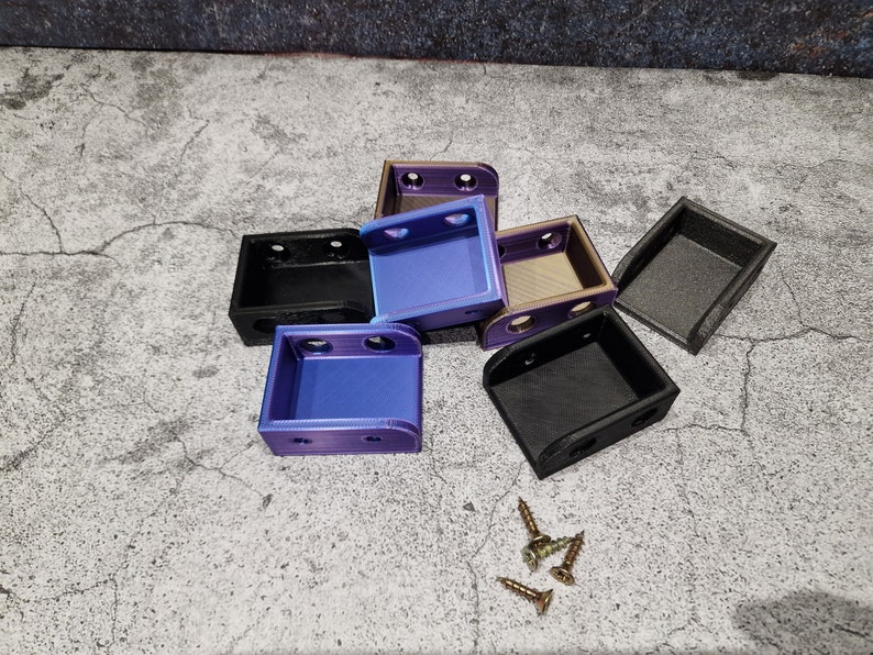May include: Six small, 3D printed boxes in various colors, including black, purple, and gold. The boxes are stacked on top of each other. There are four small screws on the surface.
