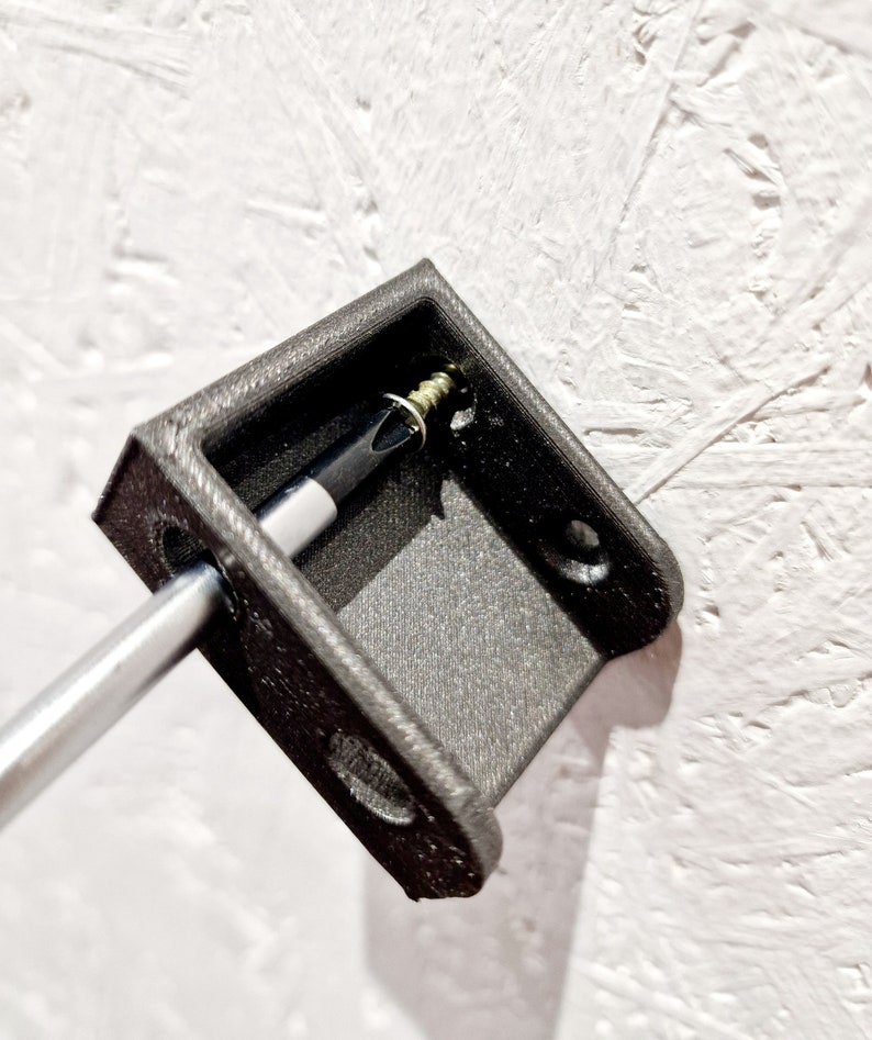 May include: A black plastic wall mount with a screw inserted into it. A screwdriver is being used to tighten the screw.