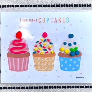 May include: A white card with the text "I can make CUPCAKES" at the top. Three colorful playdough cupcakes are shown on the card. The cupcakes are decorated with sprinkles, a swirl of pink frosting, and blueberries.