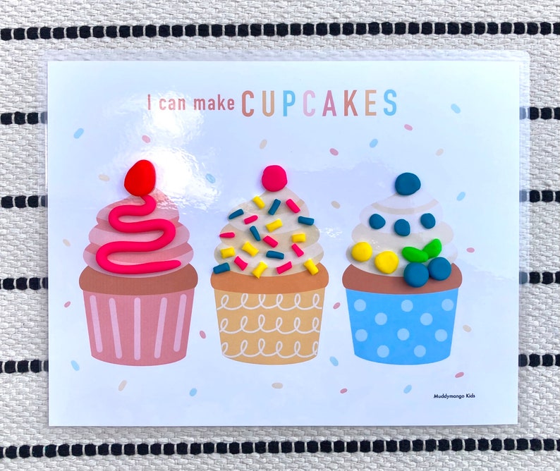 May include: A white card with the text "I can make CUPCAKES" at the top. Three colorful playdough cupcakes are shown on the card. The cupcakes are decorated with sprinkles, a swirl of pink frosting, and blueberries.
