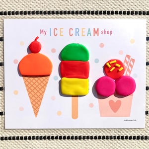 May include: A white card with the text "My ICE CREAM shop" printed on it. There are three play dough ice cream treats on the card: a red and orange ice cream cone, a green, red, and yellow popsicle, and a pink and red ice cream sundae.