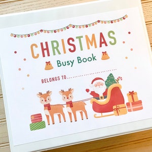 Può includere: Un raccoglitore bianco con una copertina a tema natalizio. La copertina presenta la scritta "CHRISTMAS Busy Book" con illustrazioni di Babbo Natale sulla slitta, renne, regali e una ghirlanda di luci. È visibile anche la scritta "Belongs to..."