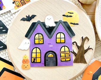 Puede incluir: Decoración de madera con temática de Halloween con una casa embrujada morada con borde negro, ventanas tapiadas y una silueta de gato negro. Incluye pegatinas de fantasmas y murciélagos, una luna llena y un árbol desnudo. Se ven pegatinas de casas adicionales.
