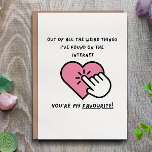 May include: A greeting card with a brown border and the text "OUT OF ALL THE WEIRD THINGS I'VE FOUND ON THE INTERNET YOU'RE MY FAVOURITE!" A pink heart with a finger pointing at it is in the center. The card is on a wooden surface.