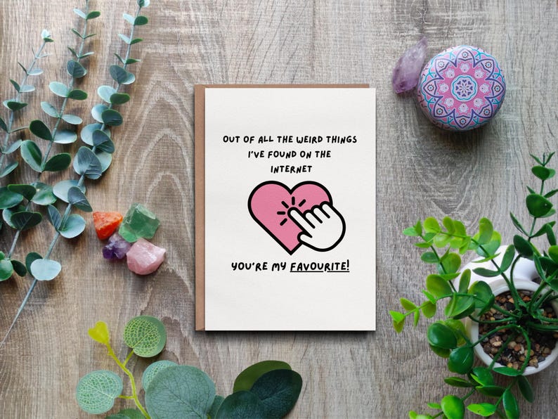 May include: A greeting card with a brown border and the text "OUT OF ALL THE WEIRD THINGS I'VE FOUND ON THE INTERNET YOU'RE MY FAVOURITE!" A pink heart with a finger pointing at it is in the center. The card is on a wooden surface.