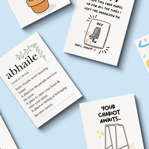 May include: A collection of greeting cards with various designs and text on a light blue background. One card features the word "abhaile" with an Irish definition. Other cards have illustrations of a light switch, a cake, and a walker.