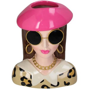 May include: A ceramic planter shaped like a woman's bust. The woman is wearing a pink beret, large round sunglasses, gold hoop earrings, a gold chain necklace, and a leopard print top.