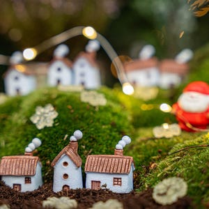 Può includere: Scena di un villaggio natalizio in miniatura con case bianche, tetti marroni e camini fumanti. Sono presenti anche una figura di Babbo Natale e un albero di Natale verde. La scena è ambientata su muschio verde, con luci a corda.
